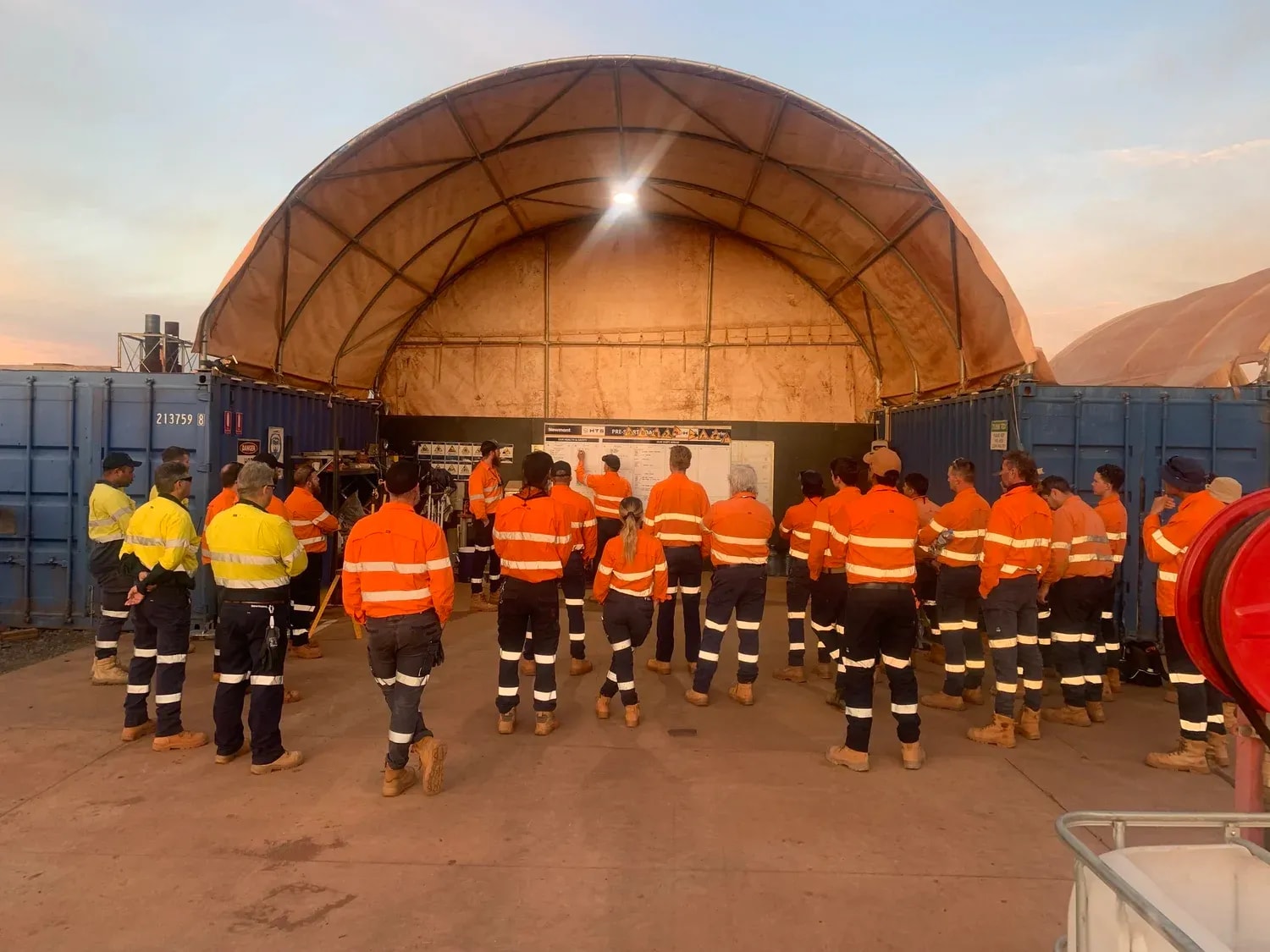 Crew gathered for a pre-start meeting under a shelter at a remote site at dawn