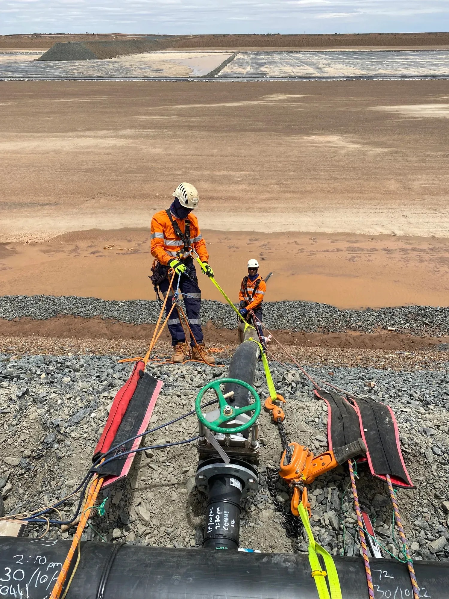 Two workers in helmets and hi-vis rigging a pipeline section beside a tailings dam